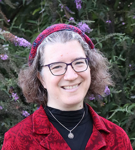 Image description: A photograph of Julia Watts Belser, a white Jewish woman with curly brown hair. She's wearing a patterned red blazer and a red kippah (beret). In the background is a tree with purple flowers.