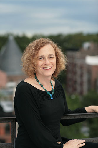 Image description: A photograph of Joy Ladin, a white Jewish woman with curly blonde hair. She's wearing a black shirt and turquoise necklace. In the background is a blurred cityscape.